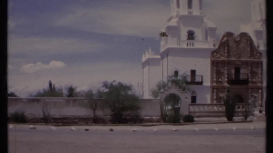 1971 California 8mm frame showing a white religious building with twin towers, arched entry, low wall, desert shrubs, and paved road.
