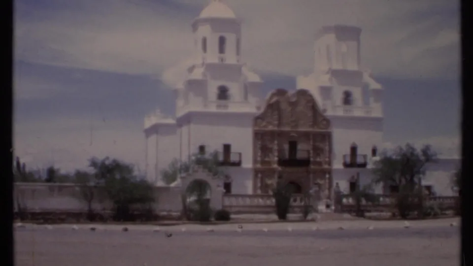 1971 California home-movie view of a white church or cathedral facade with towers, arched entry, roadside pavement, and sparse vegetation.