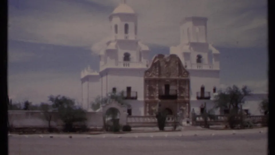 1971 California 8mm frame with a closer roadside view of the church towers, decorative facade, low wall, shrubs, and open sky.