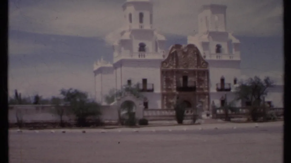 1971 California home-movie frame showing the white church, ornate central entrance, towers, roadside pavement, low wall, and arid landscaping.