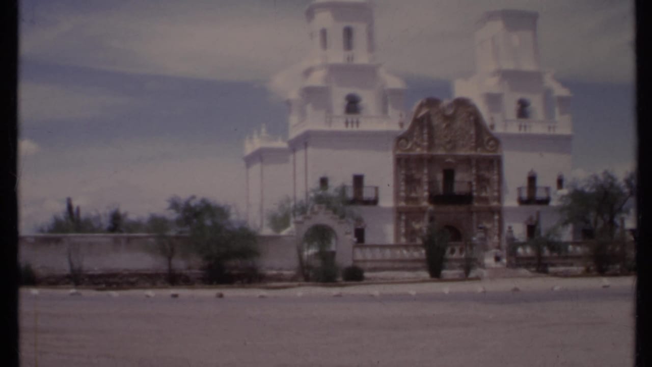 White church exterior with twin towers, ornate yellow entrance, low wall, and desert plants.