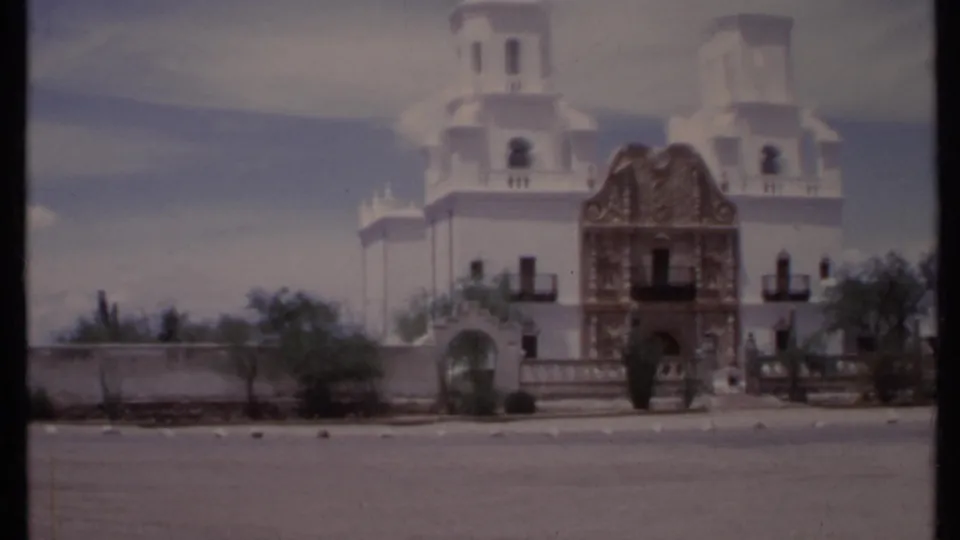 1971 California 8mm frame showing a white church with twin towers, ornate yellow entrance, roadside foreground, and sparse desert landscaping.