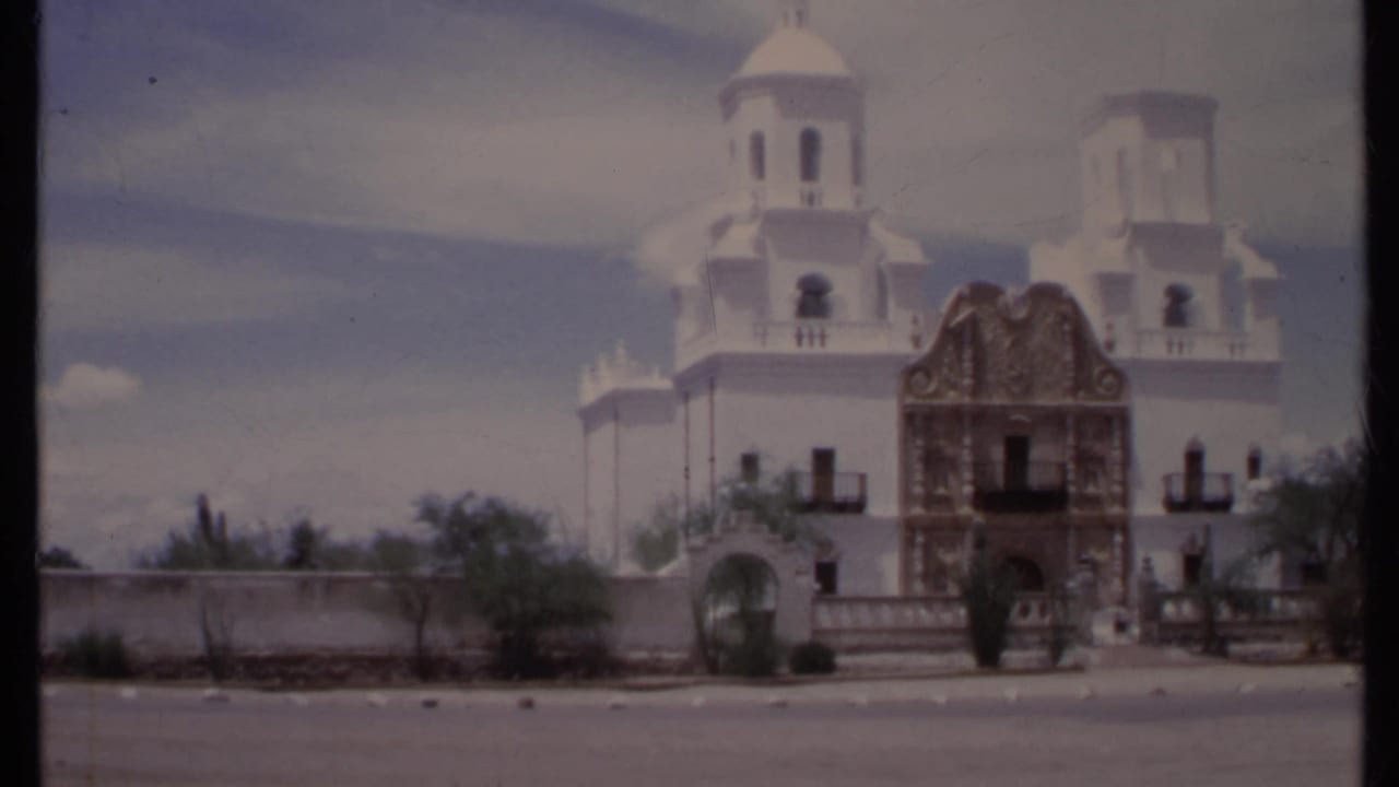 White church with bell towers and decorative entrance behind a low wall and roadside.
