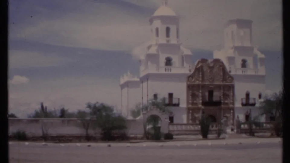 1971 California home-movie frame of a white church exterior, twin towers, arched side gate, decorative yellow entrance, and desert plants.