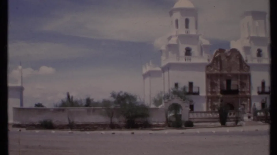 1971 California frame with a softly focused white church, twin tower forms, ornate entrance, low wall, shrubs, and paved road in front.
