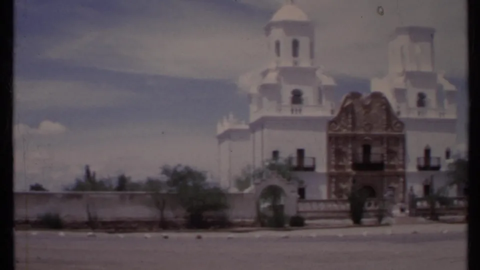 1971 California 8mm frame showing the church front from across a road, with twin towers, ornate entrance, low wall, and desert planting.