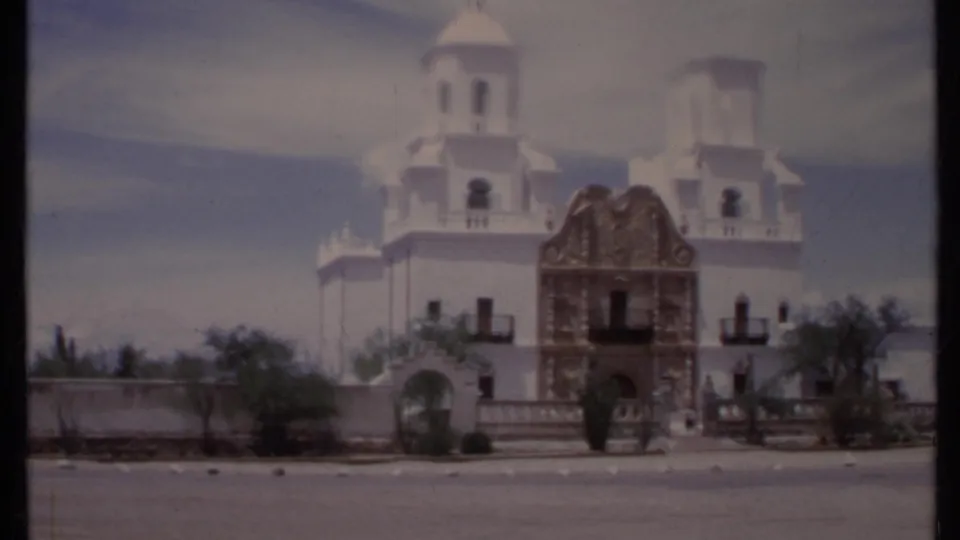 1971 California 8mm view of a white mission-style building with brown stonework, twin towers, low wall, and street foreground.