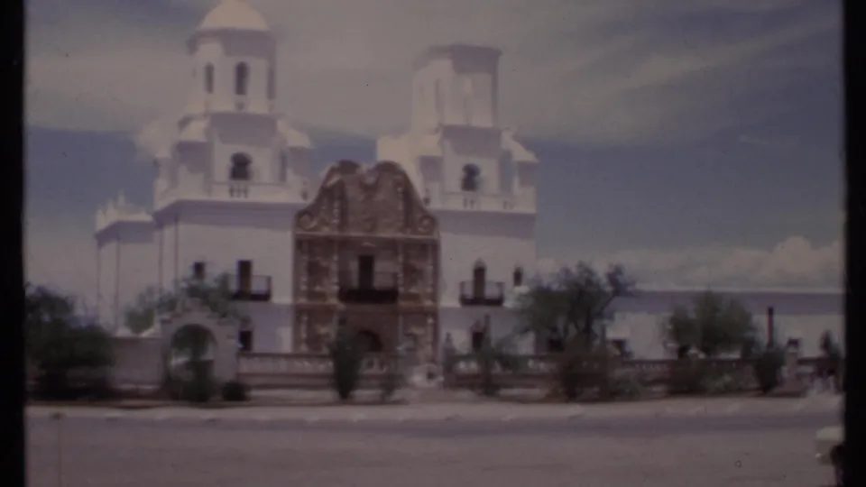 1971 California home-movie frame showing the mission-style exterior, arched entrance, garden plantings, and roadway in front.