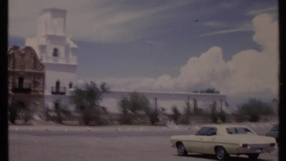 1971 California home-movie view of the building edge, low wall, small visitors, desert landscaping, and a pale green sedan on the street.