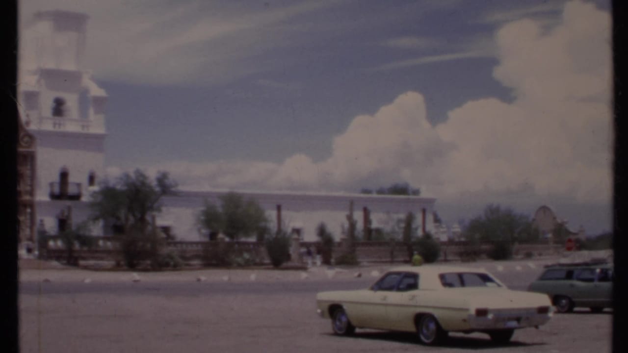 White building and parked cars beside an open lot under a cloudy blue sky.