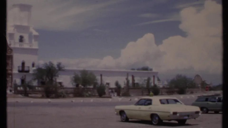 1971 California 8mm frame showing a white building, several parked cars, desert plants, and low mountains beyond the lot.
