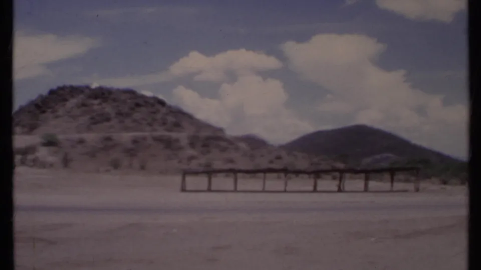 1971 California view of a rocky desert hill, open paved foreground, low horizontal structure, and bright clouds.