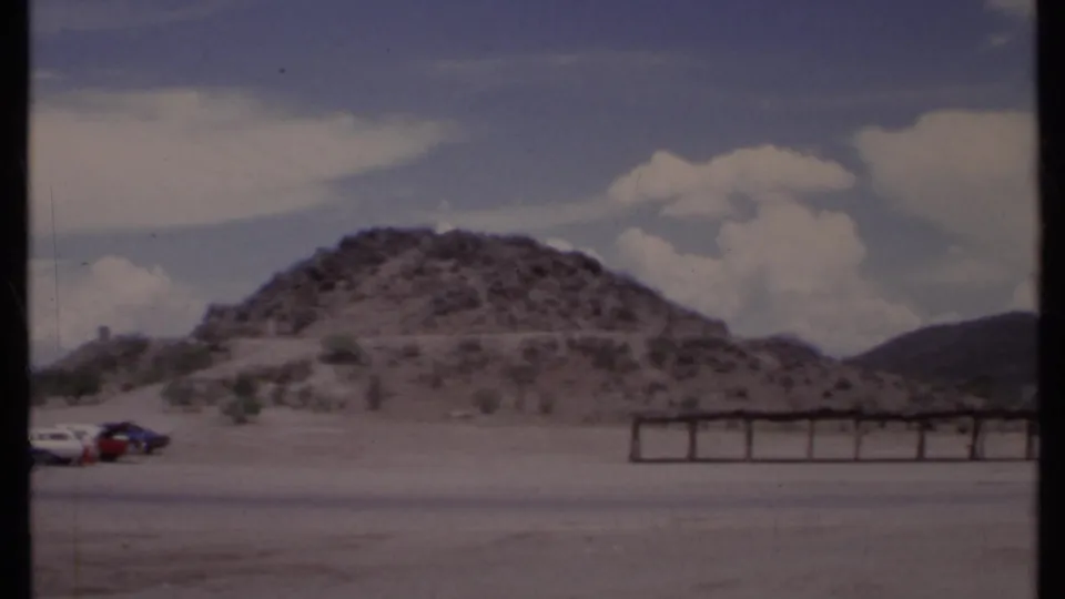 1971 California frame showing a rocky hill, parked cars at the edge of the lot, desert plants, and cloud-filled sky.