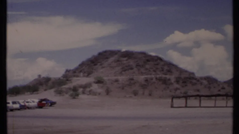1971 California 8mm frame of a broad paved foreground, low desert hill, parked cars, and a simple horizontal structure.