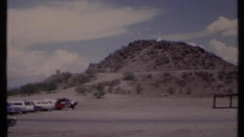 1971 California 8mm frame of a dry hillside, parked cars, sparse shrubs, and a small wooden shelter.