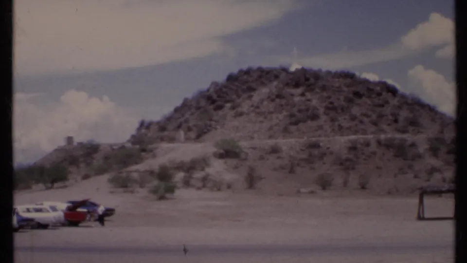 1971 California home-movie frame zooming toward a rocky hill with a white cross visible near the top.