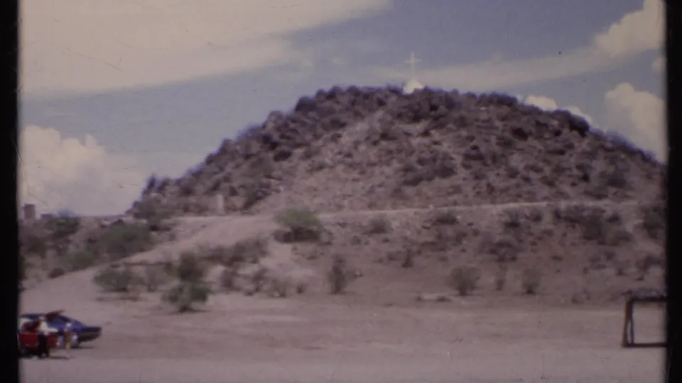 Closer 1971 California 8mm view of a dry rocky hill with a white cross and sparse desert vegetation.