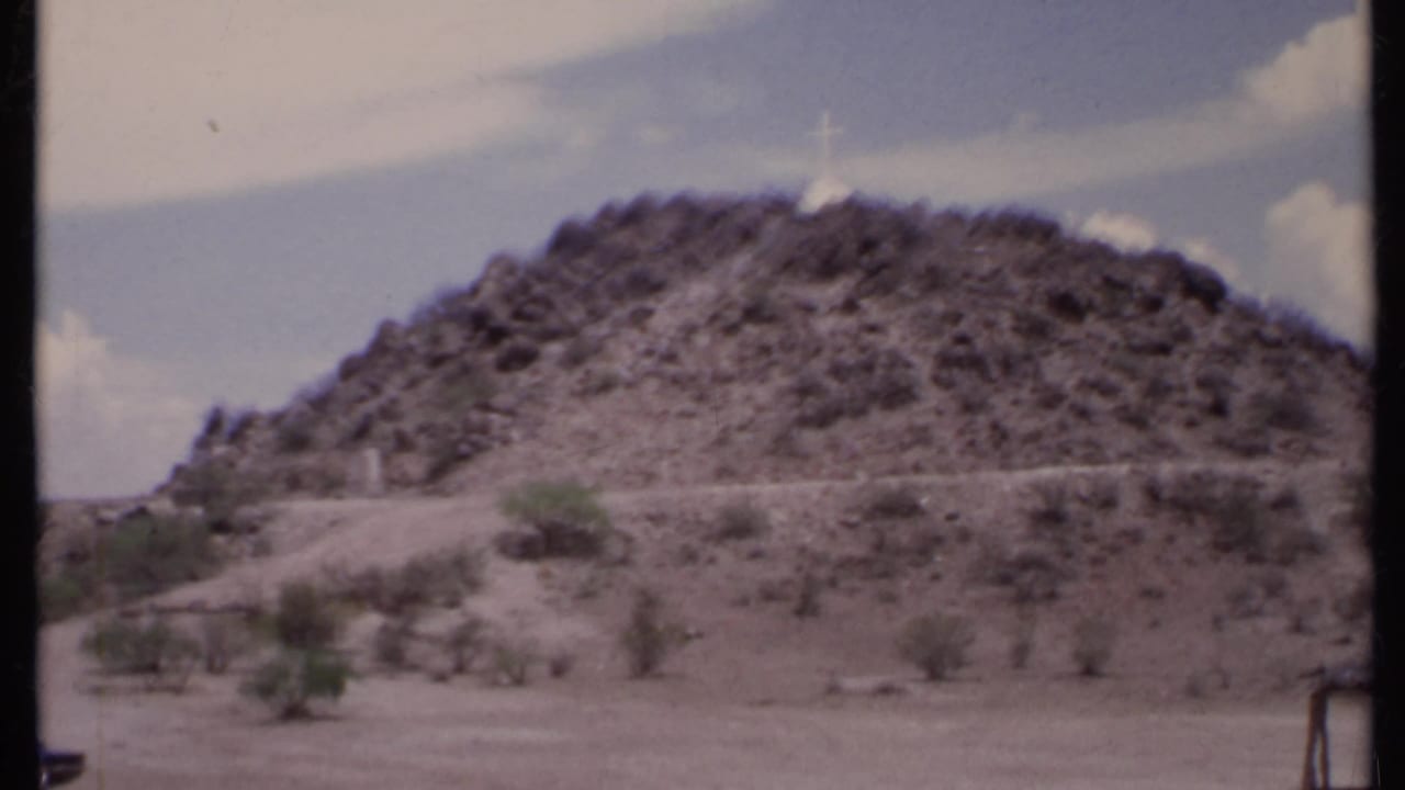 Rocky desert hill with a white cross on the peak under a pale sky.