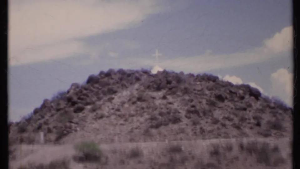 Soft 8mm view of a California desert hill in 1971, with scrub at the base and a white cross near the rocky crest.