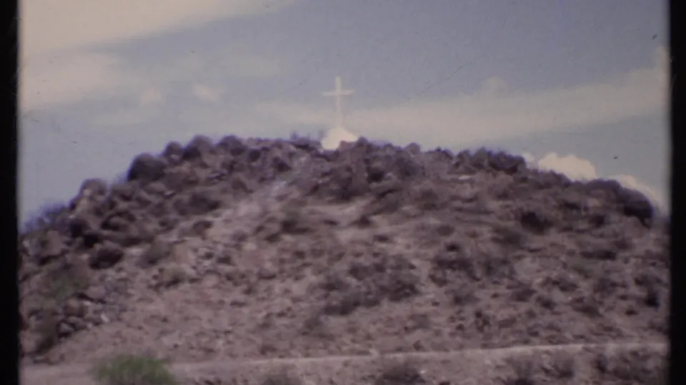 1971 California home-movie frame with the cross more prominent above a rocky, scrub-covered desert slope.