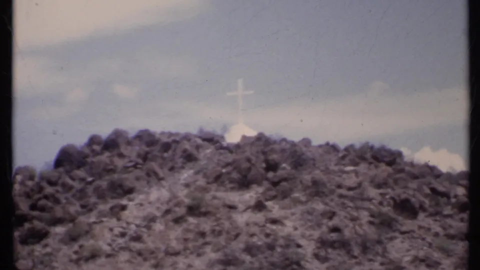 Soft 1971 8mm frame of a California desert hilltop cross, with rocky terrain and bright sky filling the view.