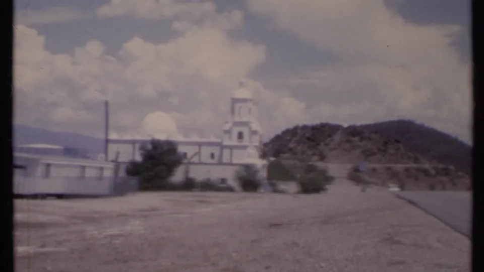 1971 California 8mm frame of a white hillside church and bell tower seen from an open dirt area.