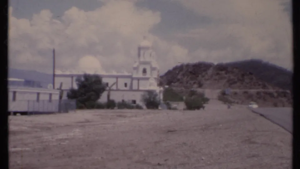 1971 California 8mm frame showing a white church with bell tower and dome near a desert road and rocky hillside.