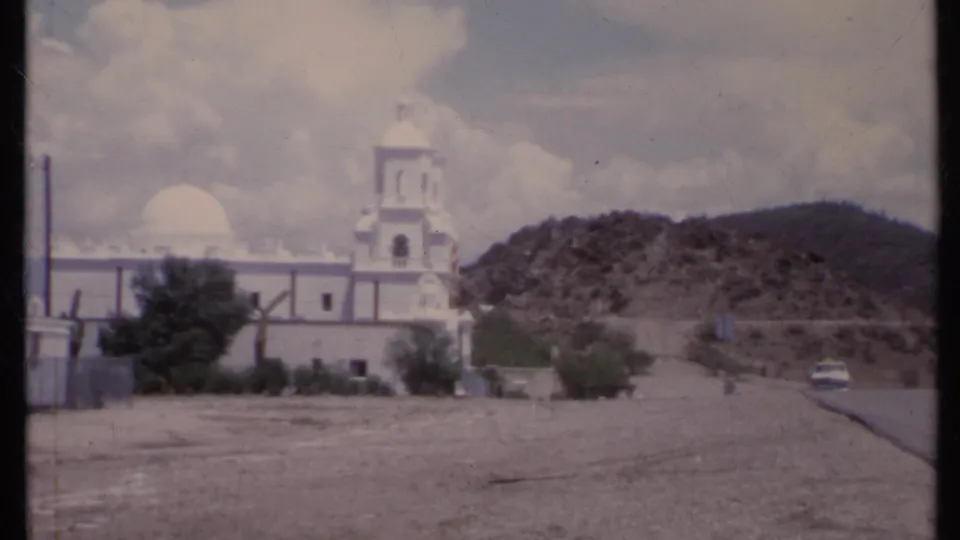 1971 California 8mm frame of a white church beside a desert road, with a car near the right edge and hills behind.
