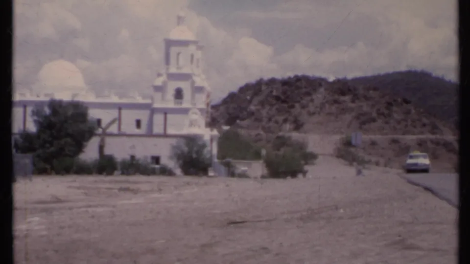 1971 California 8mm frame with a closer view of the church facade, dome, bell tower, desert shrubs, and passing car.