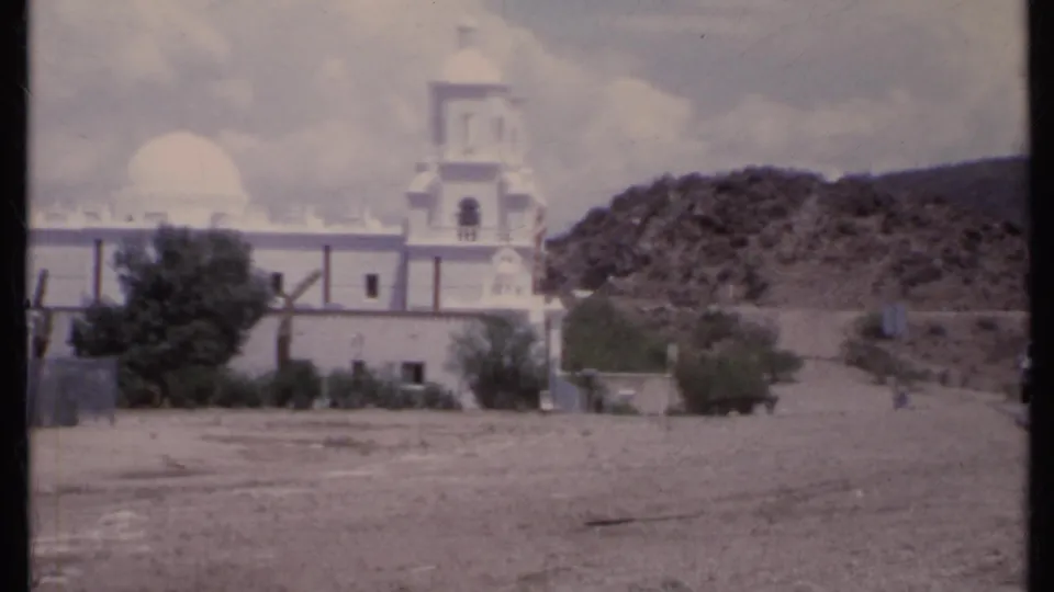 1971 California 8mm frame showing the white church and bell tower against a rocky desert hillside under a partly cloudy sky.