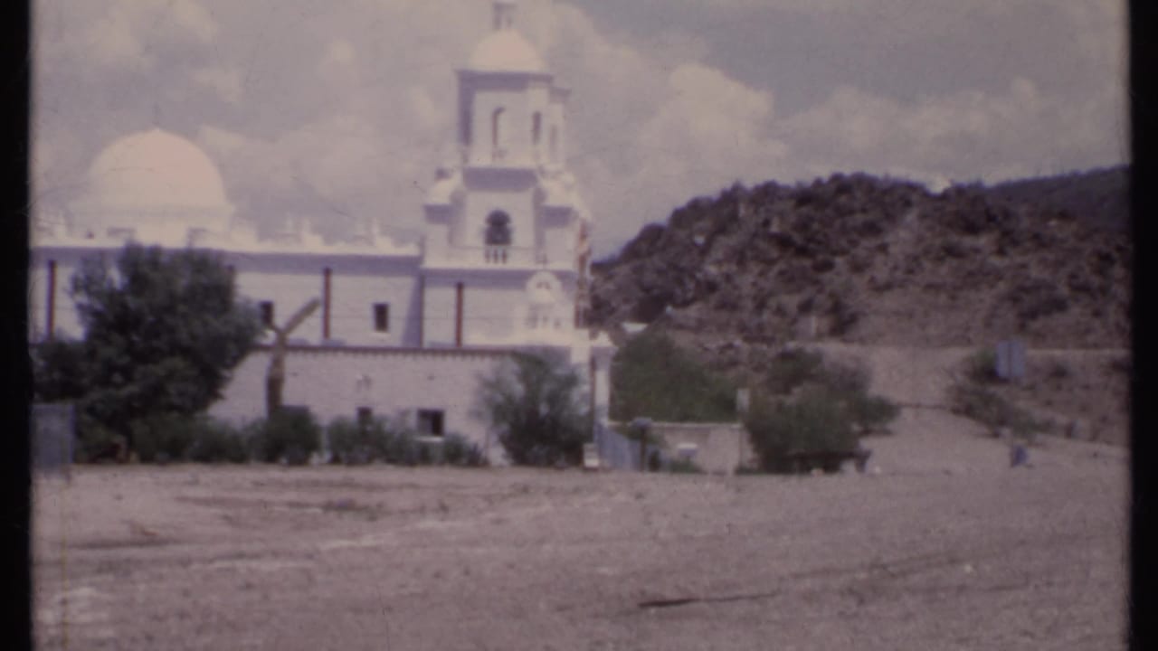 White church with dome and bell tower beside a rocky hillside and open dirt foreground.