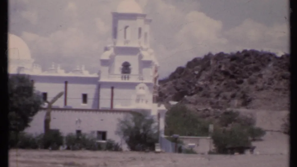 Closer 1971 California 8mm view of the white church tower and dome edge with desert shrubs and rocky hillside behind.