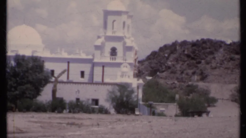 1971 California 8mm frame showing the church, bell tower, dome, desert hillside, shrubs, and a small vehicle near the road.