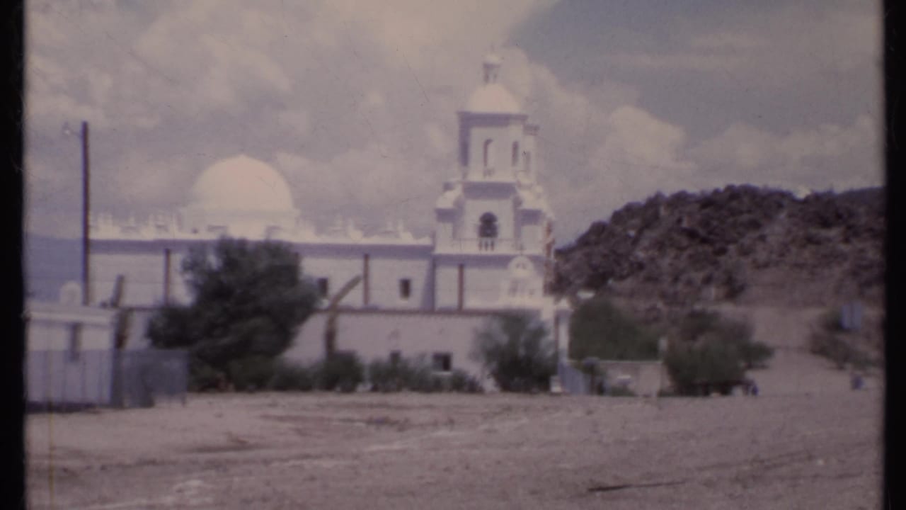 White church with bell tower and dome beyond a dirt field, with trees and a rocky hill nearby.