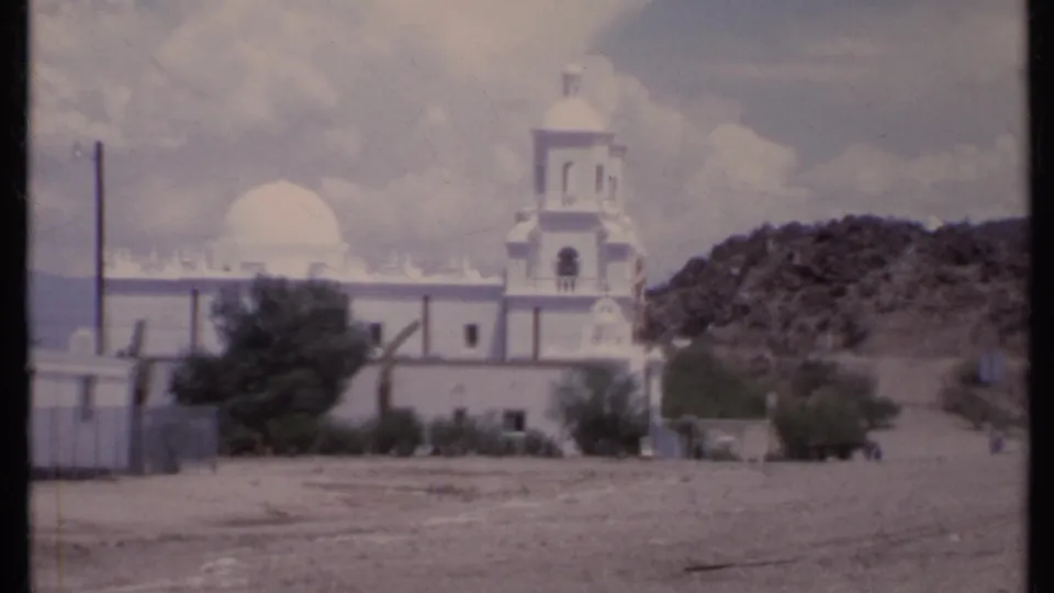 1971 California 8mm frame showing a white church with bell tower and dome across a dusty open lot, with scrub trees and hillside behind it.