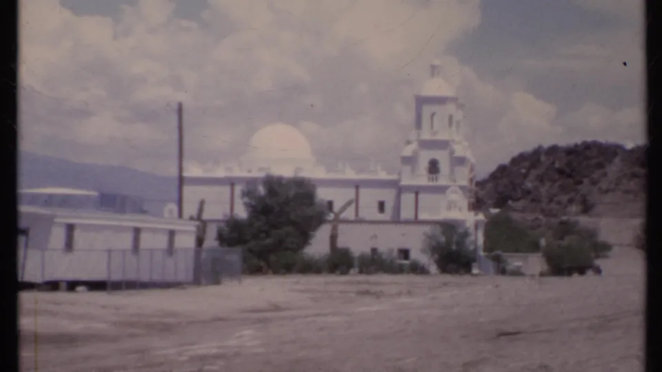 1971 California home-movie frame with a white church partly framed by a mobile home, utility pole, dirt foreground, and low hillside.