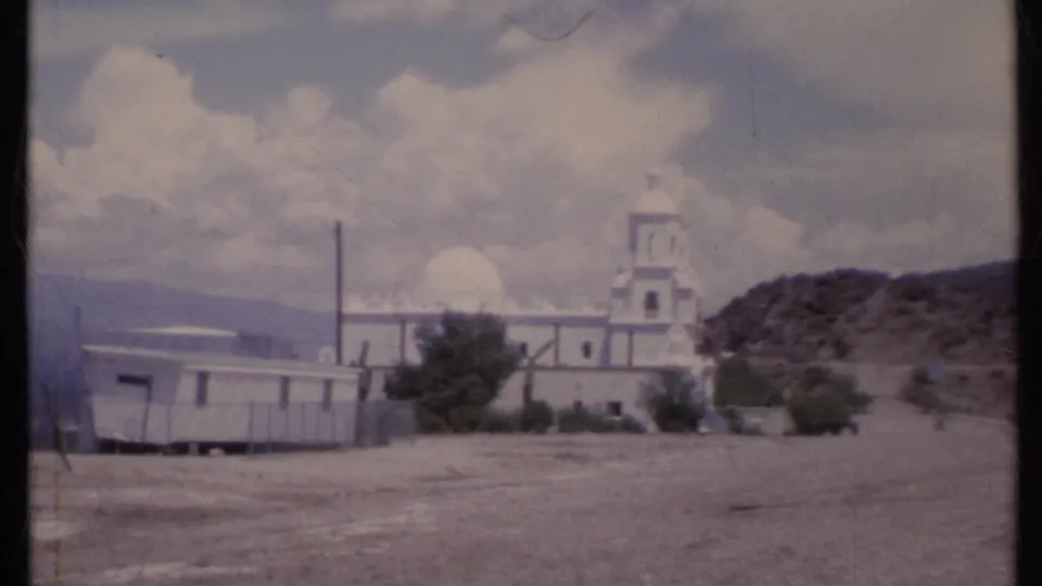 1971 California 8mm view of a white church receding in frame, with a mobile home, utility pole, dry ground, and mountains in the background.