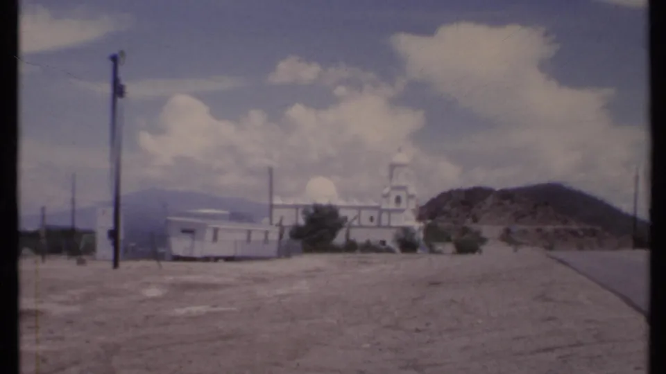 Wide 1971 California home-movie frame showing the church small in the distance across an open dirt field, with poles, hillside, and partly cloudy sky.