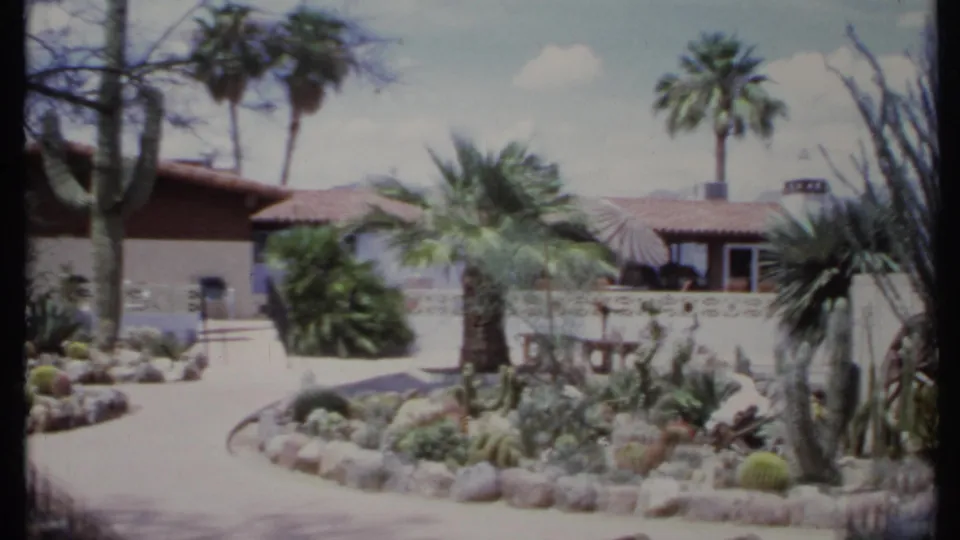1971 California home-movie view of a curved driveway, cactus garden, palms, stucco walls, and terracotta roof under bright sun.
