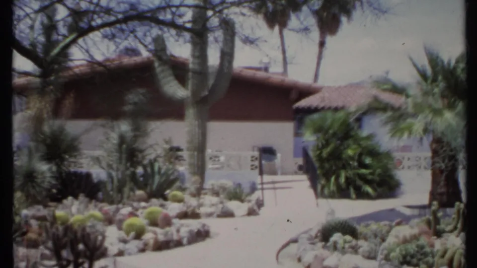 1971 California home-movie view of a stucco residence with a tall cactus, palms, red tile roof, and desert planting beds.