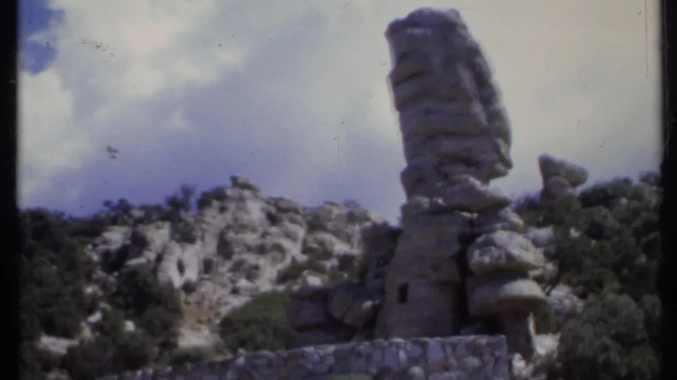 1971 California outdoor landscape with tall cactus, low stone wall, scrubby hillside, pale rocks, and cloudy sky on 8mm film.