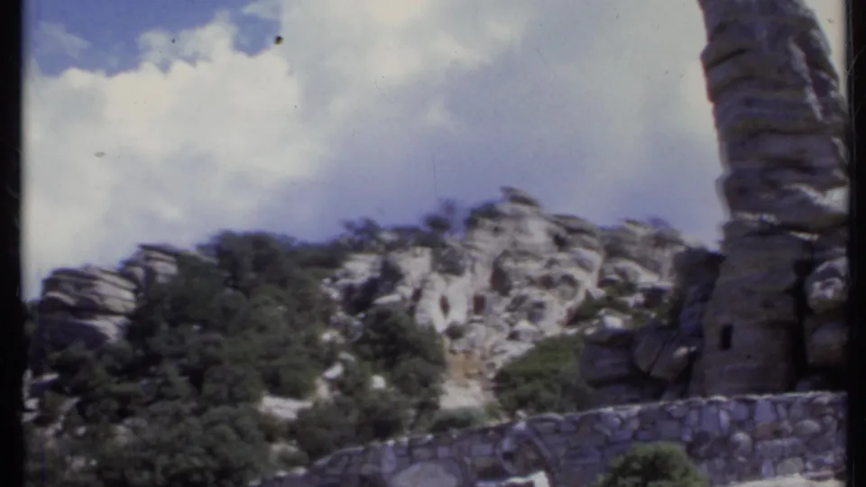 1971 California landscape detail showing a stone wall, scrubby rock formations, cloudy sky, and a tall cactus partly at frame edge.