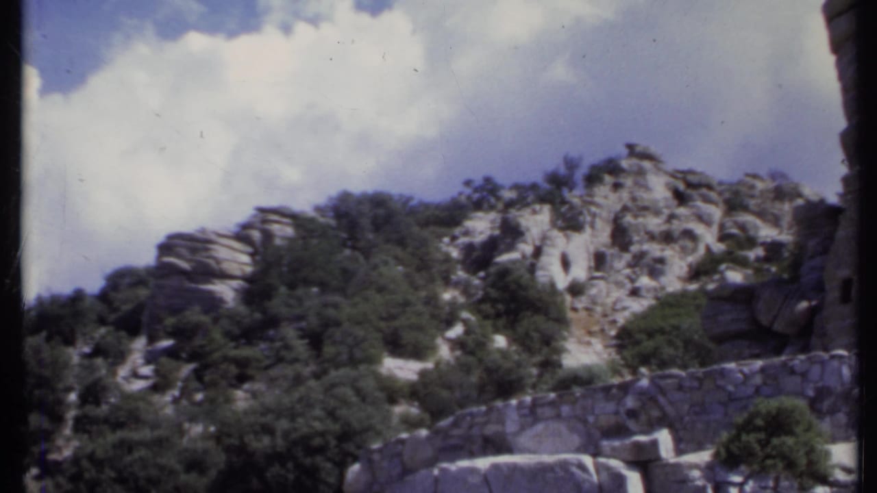 Rocky hillside with green trees, stone wall, and blue sky with clouds.