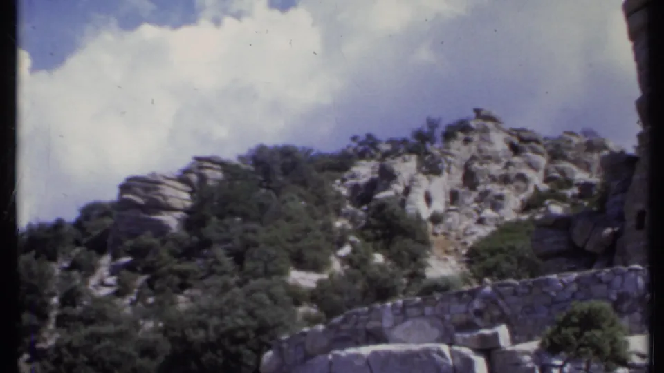 1971 California 8mm frame showing pale rock outcrops, trees, a low stone wall, and a bright clouded sky.
