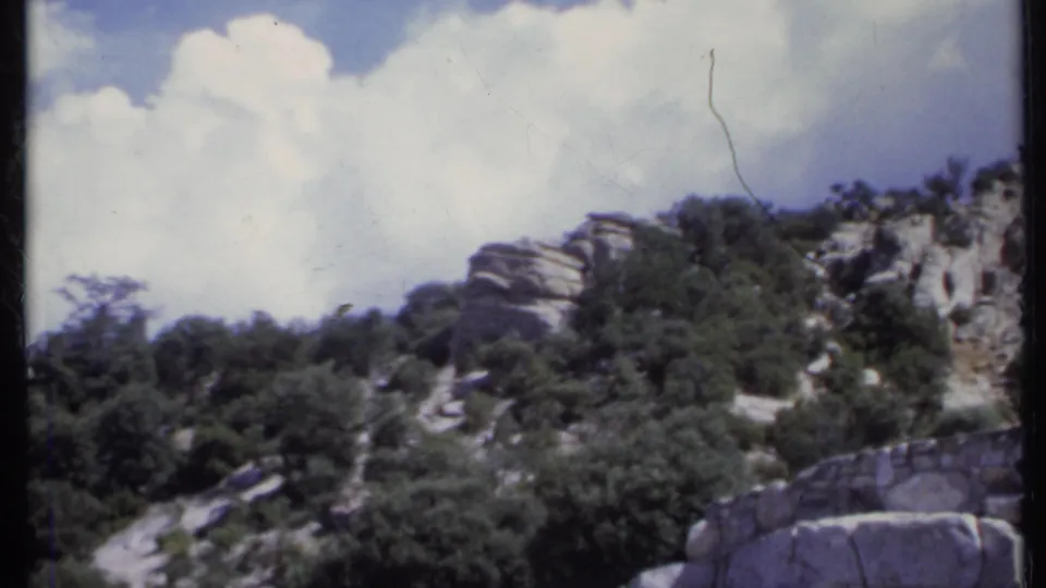 1971 California home-movie view of a rocky, tree-covered hillside with a pale path and bright clouds overhead.