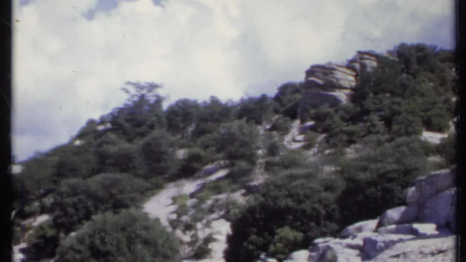 1971 California 8mm frame looking across a rocky hillside path bordered by trees and scrub vegetation.