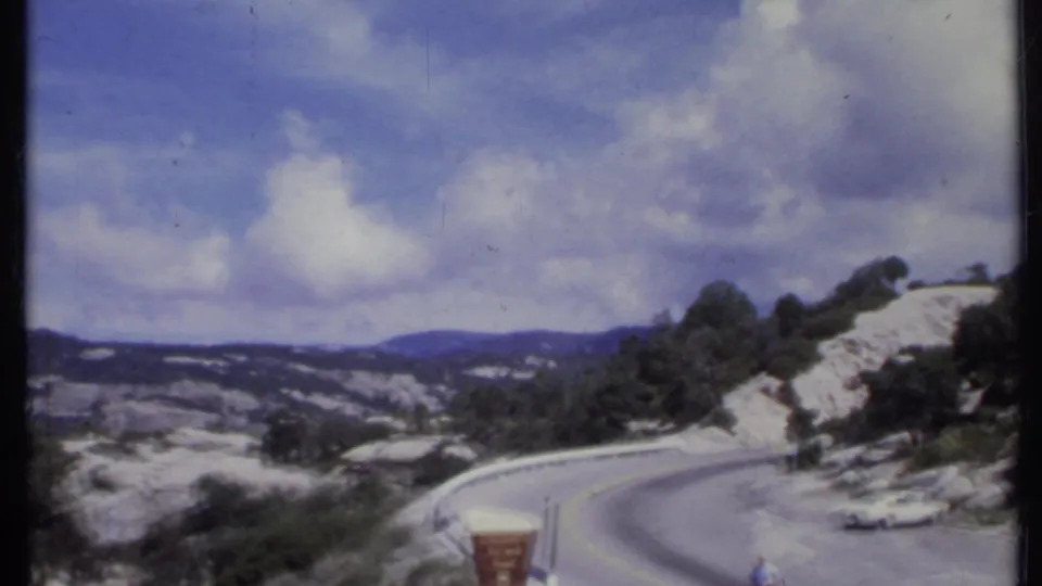 1971 California roadside view with a winding mountain road, rocky shoulder, trees, distant hills, and a small white vehicle or roadside object.