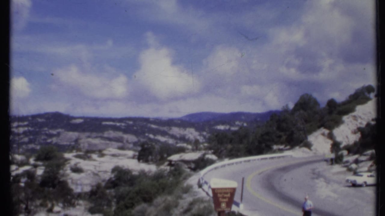 Mountain overlook with winding road, rocky hills, trees, clouds, and a roadside sign.