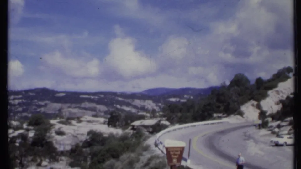 1971 California 8mm frame showing a winding mountain road beside a rocky scenic overlook with visitors and a roadside sign.