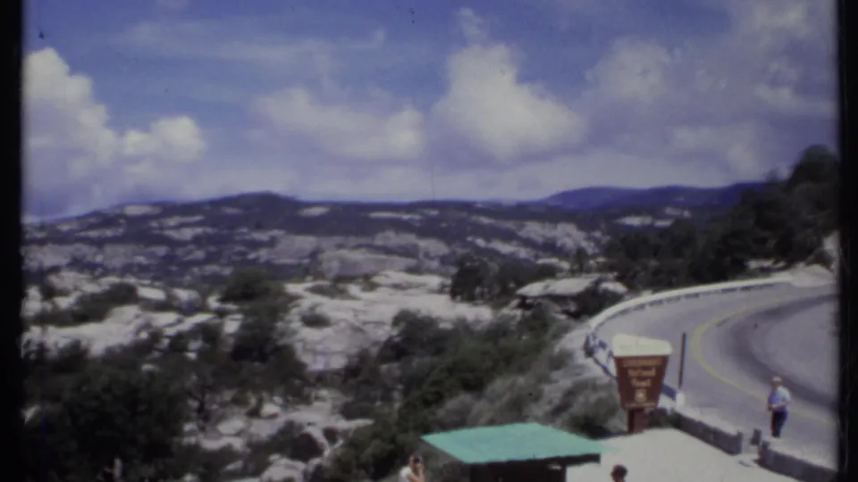 1971 California home-movie frame of visitors near a scenic overlook sign, with a parked white car and winding road above rocky hills.